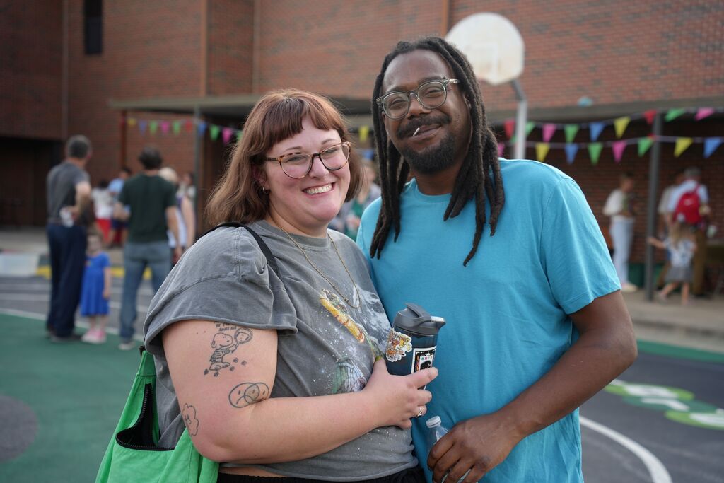 A man and woman stand in front of a building, smiling. They are surrounded by a bustling background.