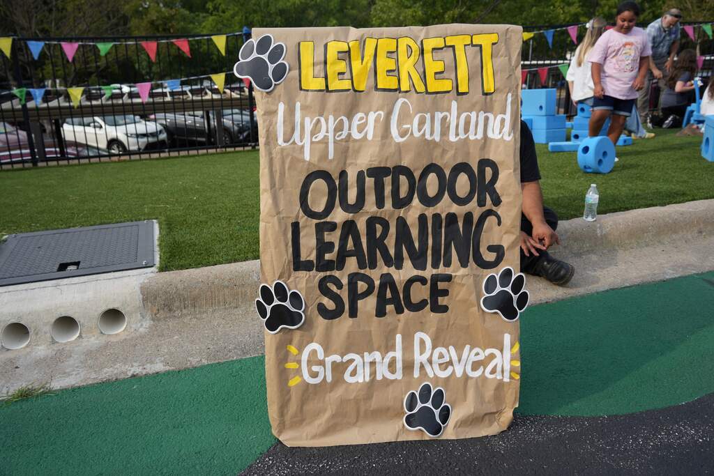 Sign in front of green grass and a sidewalk. It says "LEVERETT Upper Garland Outdoor Learning Space Grand Reveal."