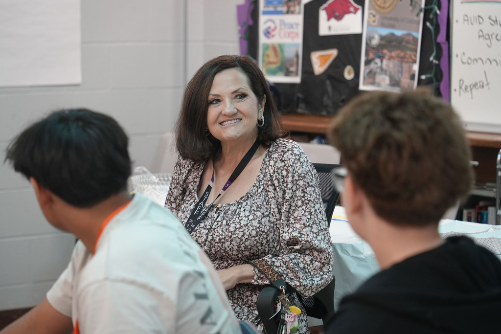A woman smiles at two students in a classroom. Behind her, a bulletin board displays various papers and pictures.