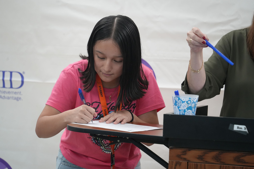 Girl in pink shirt writing on paper at a table, another person with a blue pen stands behind.