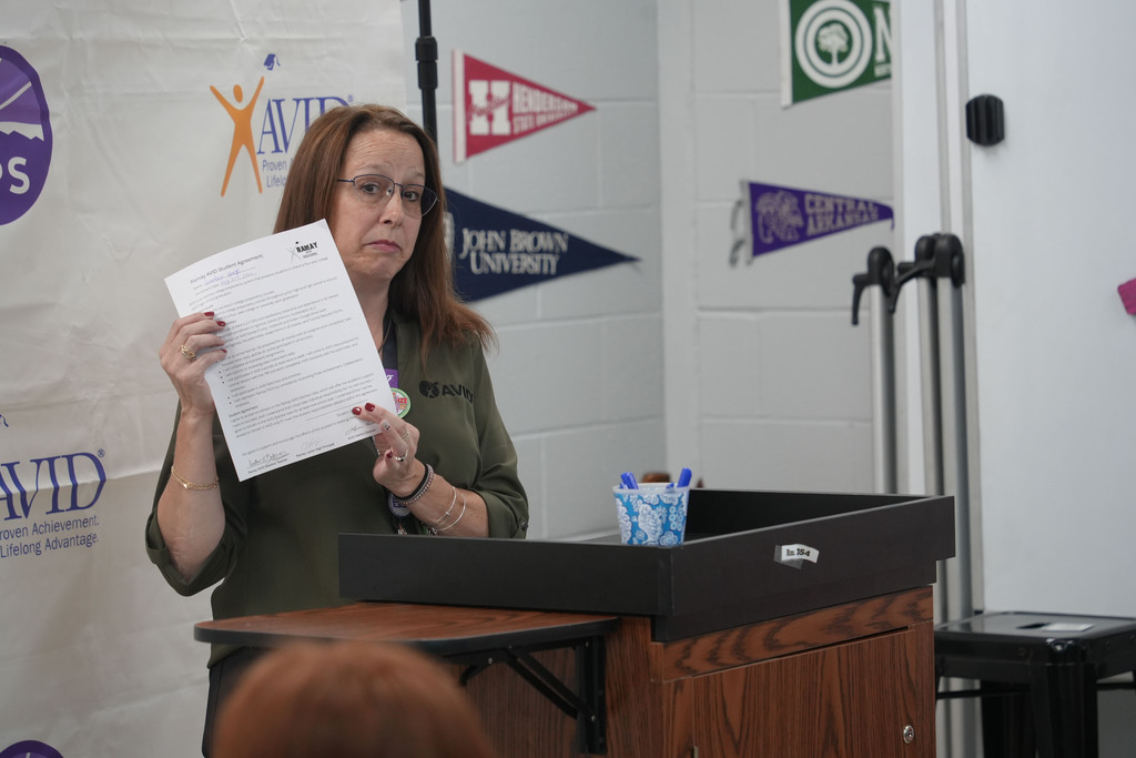 Woman with glasses standing at a podium, holding a paper, with a wall of logos and pennants behind her.