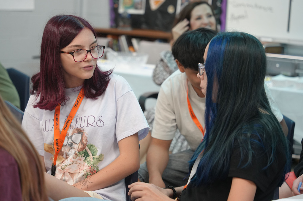 Two women, one with blue hair and the other with red, sit in a classroom and talk.