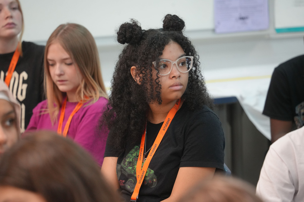 Several students sit inside a classroom. One wears glasses, a black top, and an orange lanyard.