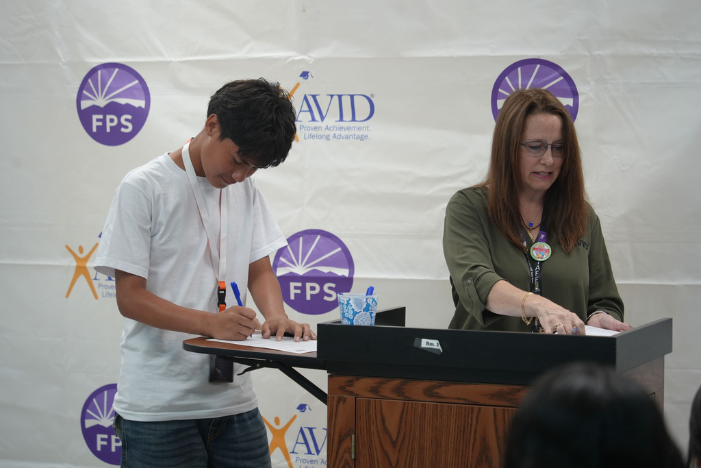 Two people at a table with a podium and backdrop. A boy writes and a woman gestures.
