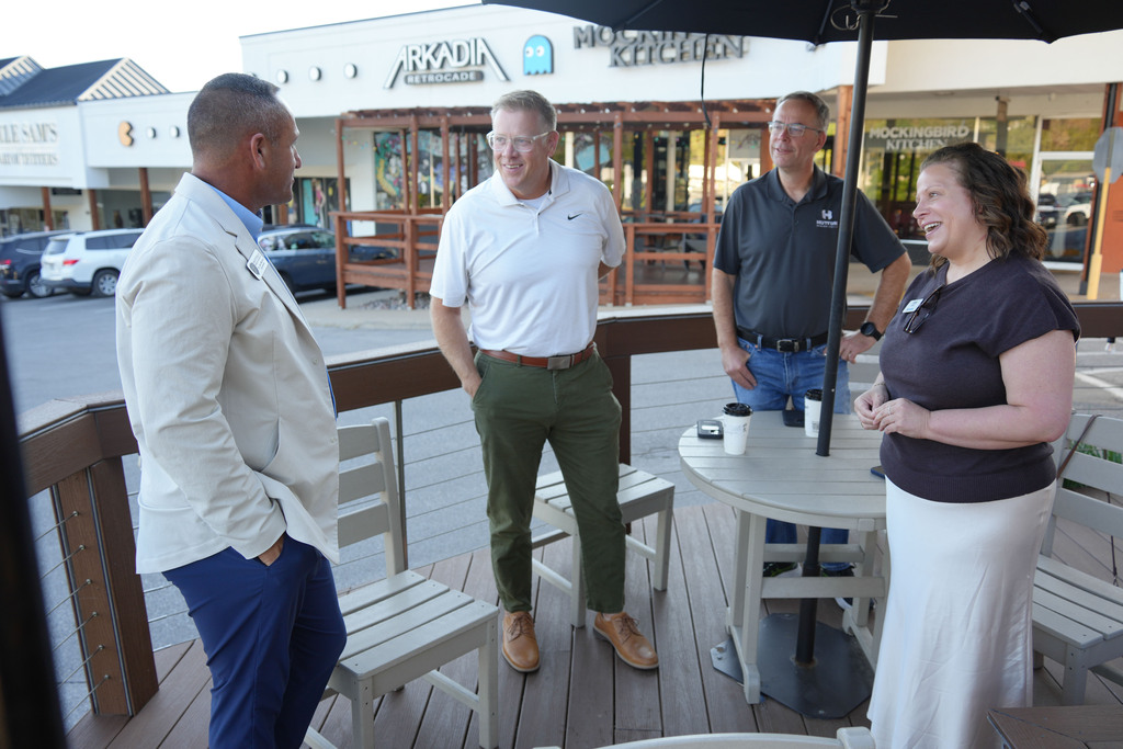 Four people stand on a deck, two men and two women, engaging in a conversation. The background shows shops.