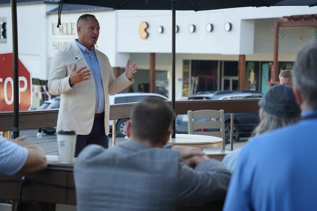 A man in a white suit gestures while speaking to a group of people seated at a table outdoors.