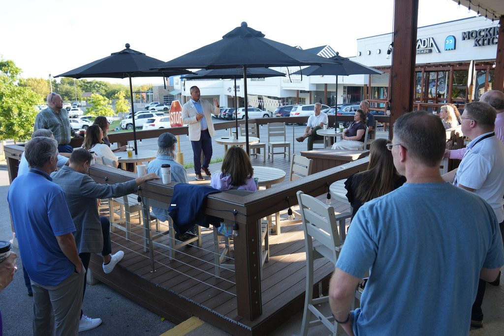People seated and standing on a patio, with a man speaking at the center. Black umbrellas provide shade.