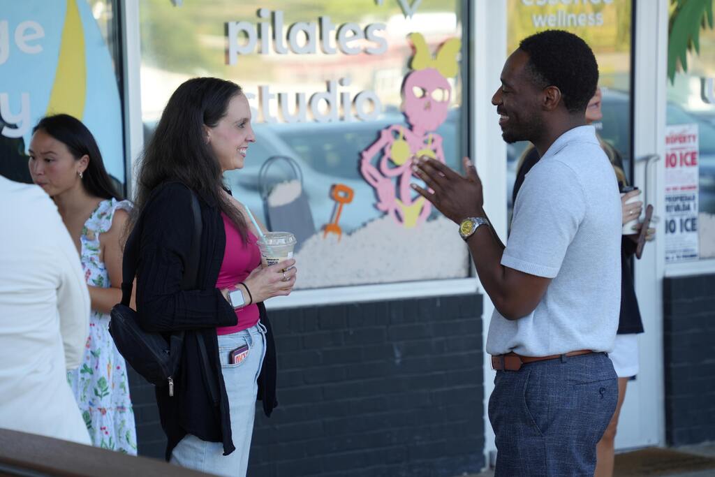 Two people converse outdoors near a sign for Pilates Studio. The woman wears a pink shirt, and the man wears a white shirt.