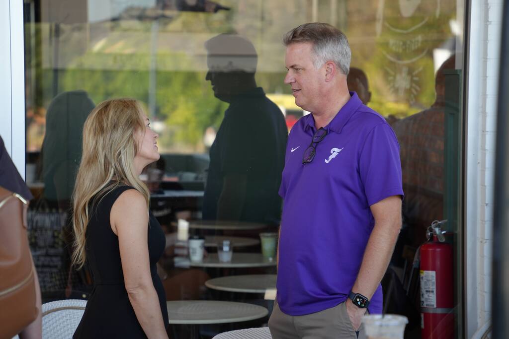 A man and woman, facing each other, talk inside a glass-walled cafe. He wears a purple polo, she a black dress.