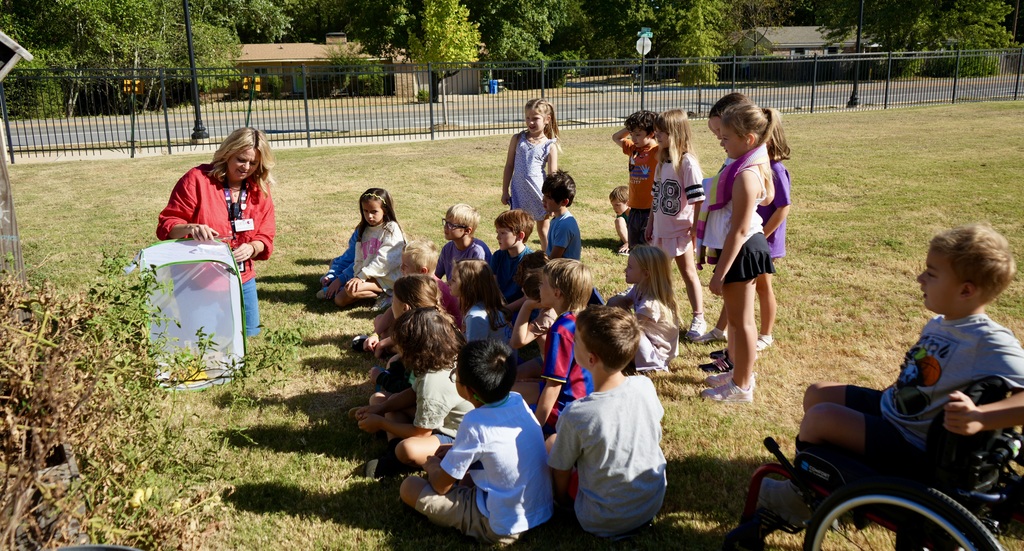An adult in a red shirt leads a group of children sitting on grass, one in a wheelchair.