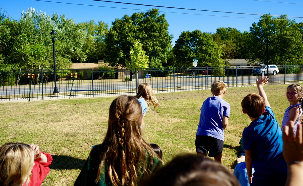 Several children stand in a grassy area near a fence, some raising their hands. Trees and a road are visible in the background.