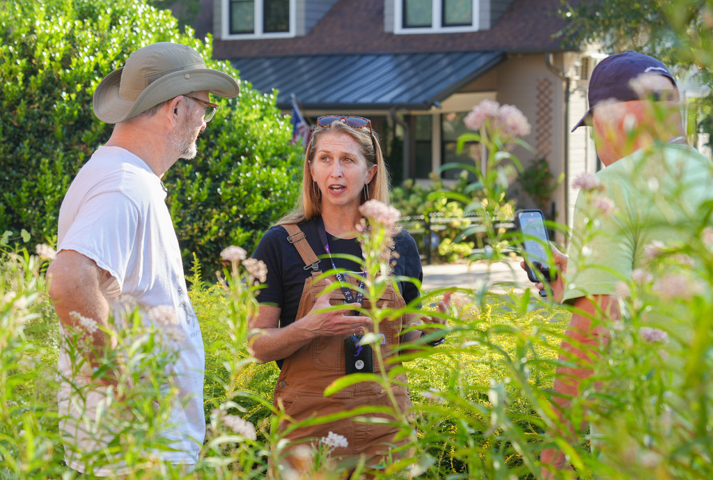 Three individuals in a garden. A woman speaks, and two men listen. House with a roof and windows in the background.