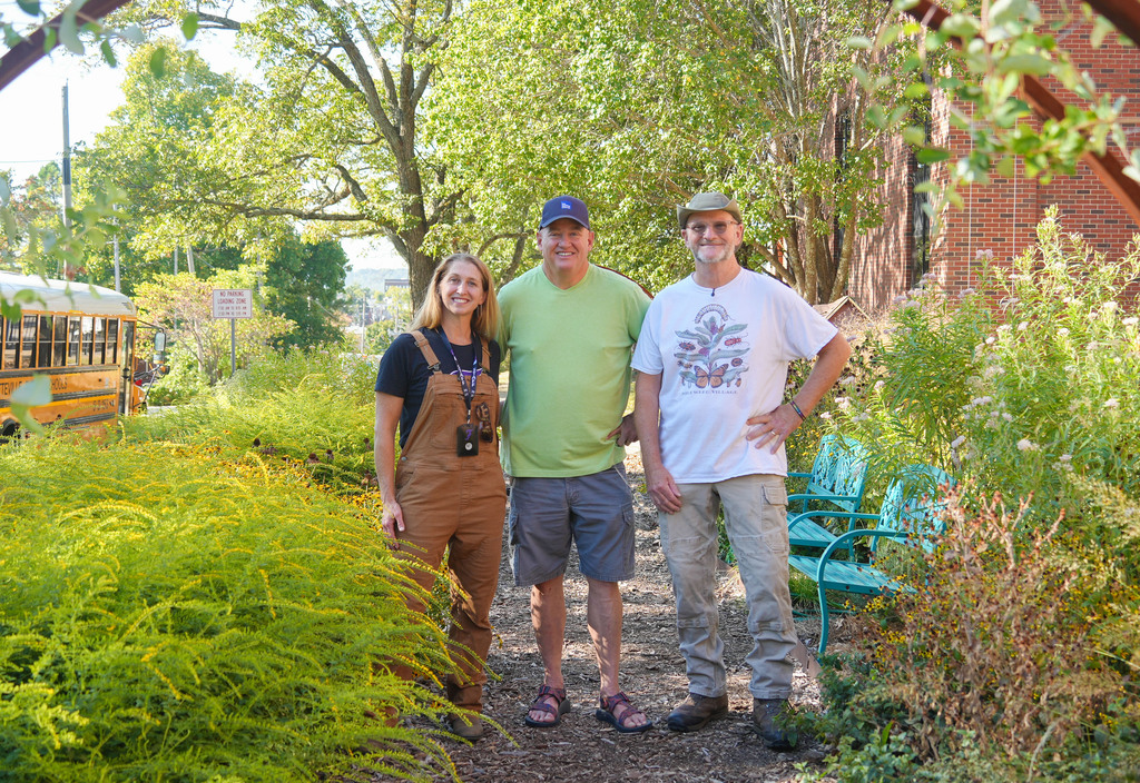 Three people stand in a garden with tall plants, blue chairs, and a bus in the background.