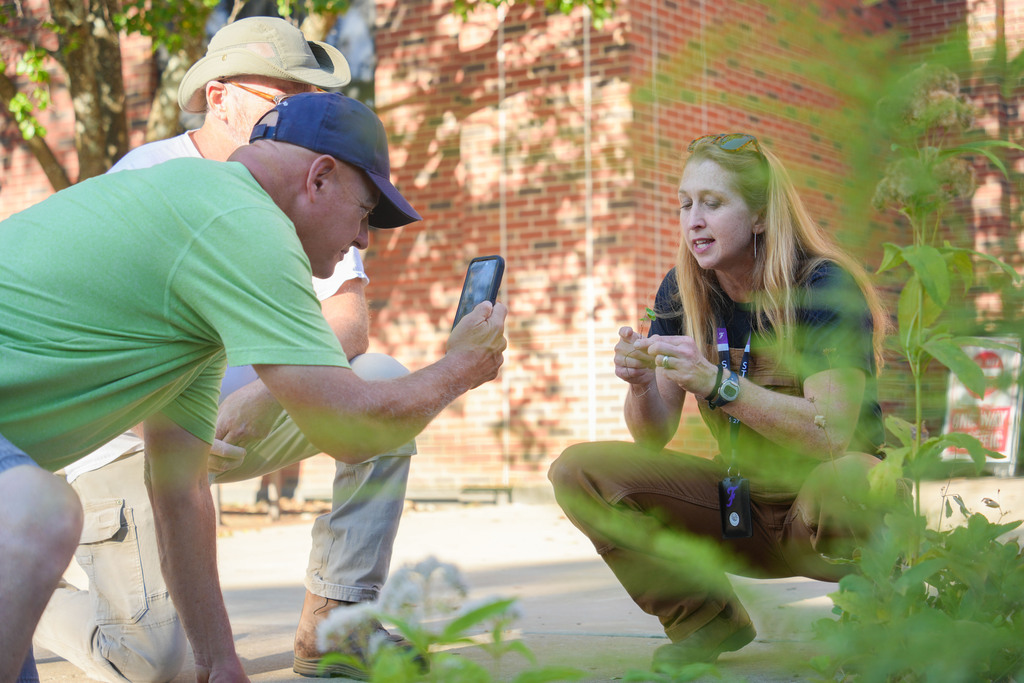 A person crouching beside another person takes a photo with a cellphone. A woman kneels nearby.