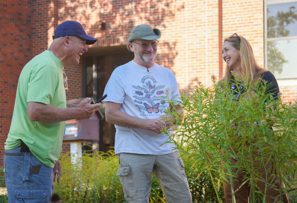 Three people near a brick building with greenery. One man shows something on his phone, another wears a cap, and a woman smiles.