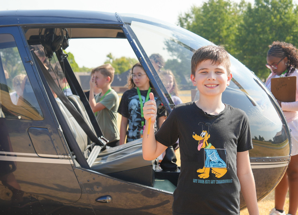 A boy stands in front of a helicopter, giving a thumbs up, with others standing behind him.