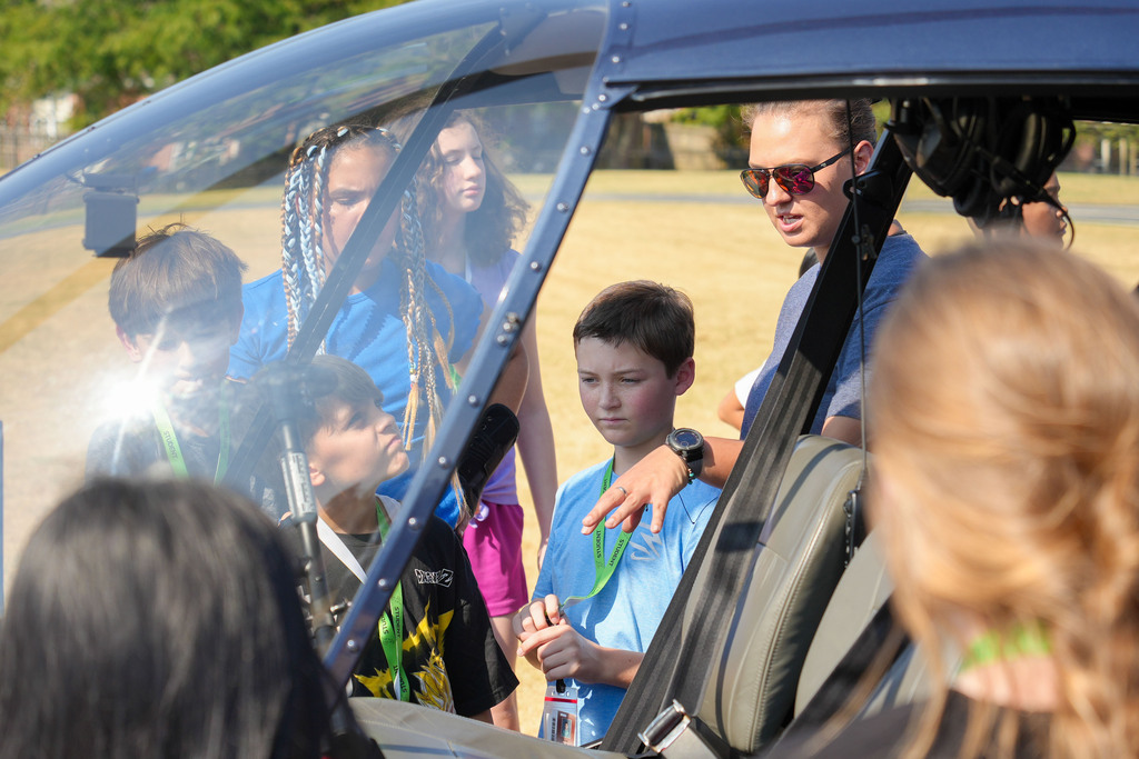 A group of children, some wearing name tags, gather around a helicopter cockpit. A man in sunglasses stands inside.