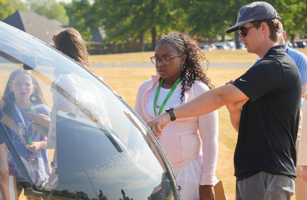 A man and a woman stand in a park, examining a vehicle. The woman wears glasses and a lanyard.