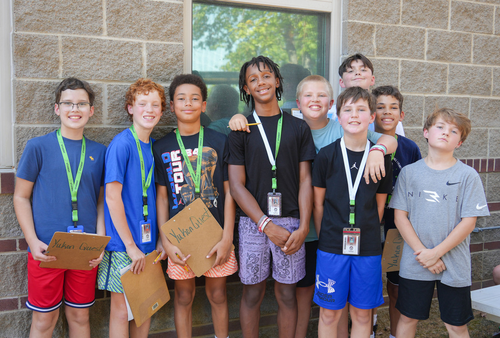 Several children in various outfits stand by a brick building, holding paper signs, and posing for a group photo.