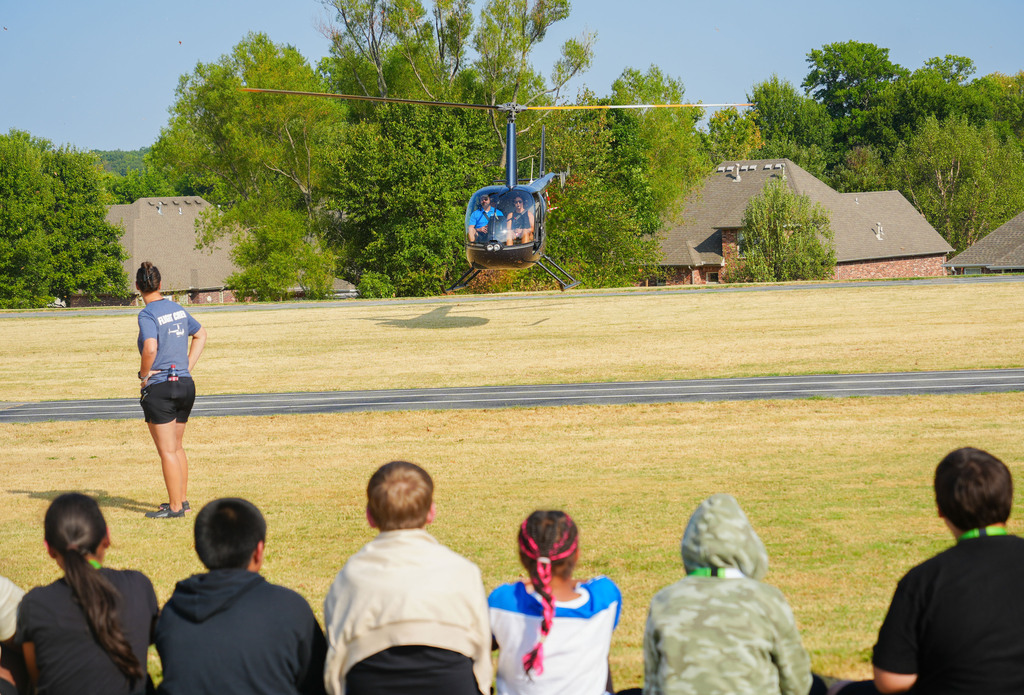 Helicopter flying over field, with people watching from the grass. Several houses in background.