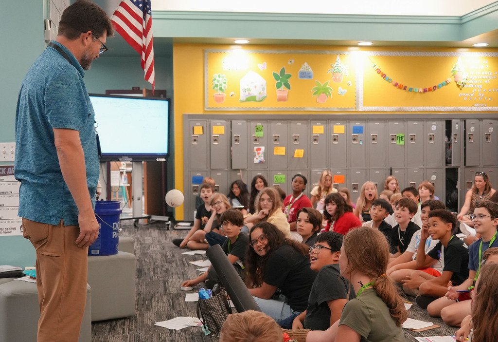 A man stands in front of a seated audience in a classroom. Lockers are behind them, and an American flag hangs on the wall.