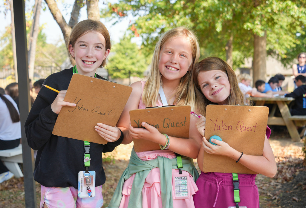 Three girls holding clipboards with written text, standing in a park. One holds a pencil, another holds a small object.