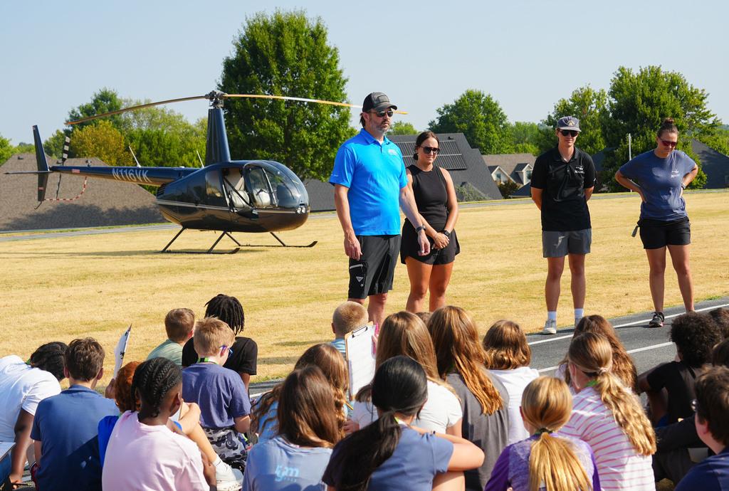 Four people stand in front of a helicopter on a field. They are addressed to a crowd of students.