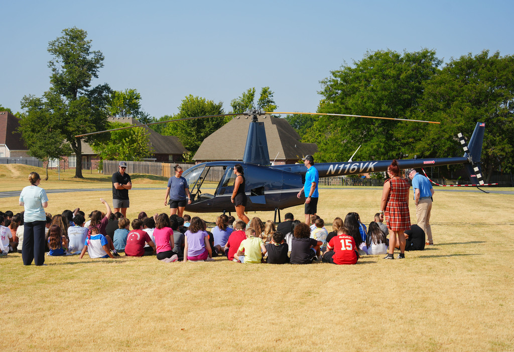 A group of people, some seated and some standing, gather around a dark blue helicopter on a field.