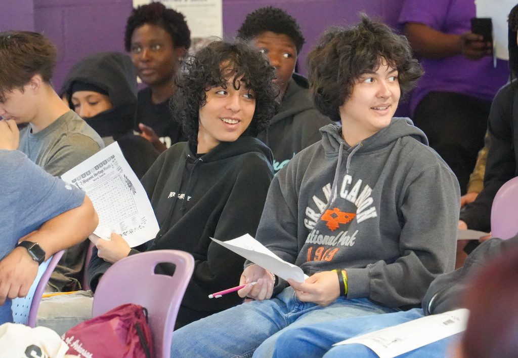 Multiple students sit in chairs, some wearing hoodies, holding papers and pens. A boy with curly hair sits next to a girl.