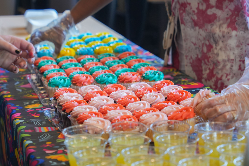 A table with colorful cupcakes and plastic cups of juice. A person wearing gloves arranges the cupcakes.
