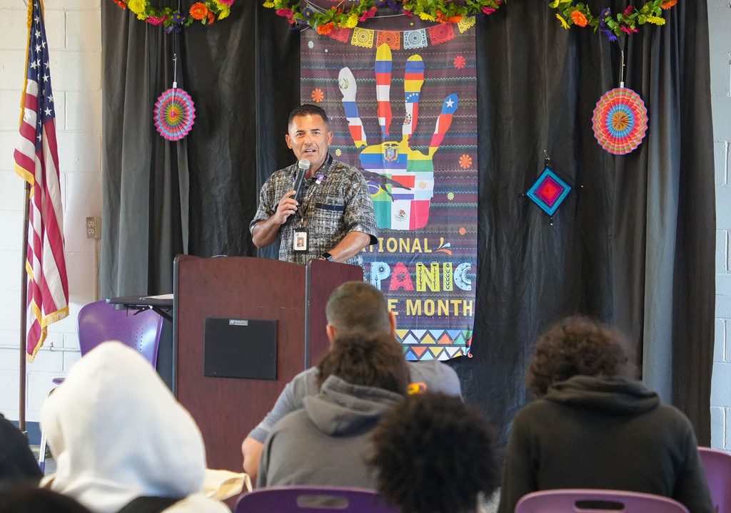 Person at podium with microphone. Behind, black curtain with colorful decorations. American flag on left. Audience of students.