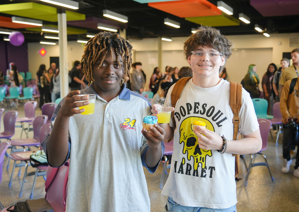 Two people hold drinks in a colorful room with chairs, balloons, and people in the background.