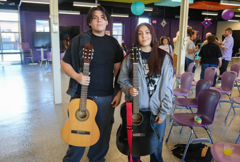 Two people hold guitars. One guitar is brown, and the other is black. They stand in a room with purple chairs.