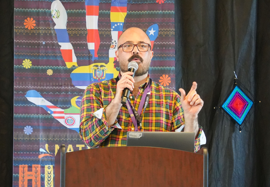 Person speaks at podium with laptop. Background displays colorful designs including a stylized hand.