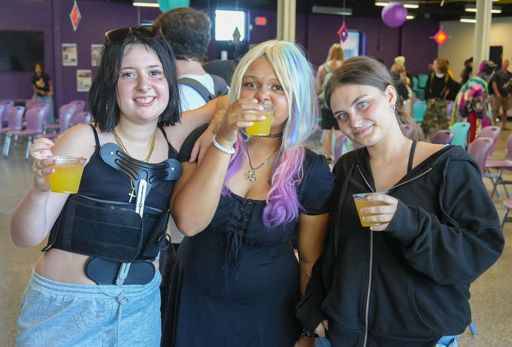 Three women pose for a photo indoors. Each holds a cup. One has colorful hair. Background includes chairs and a wall.