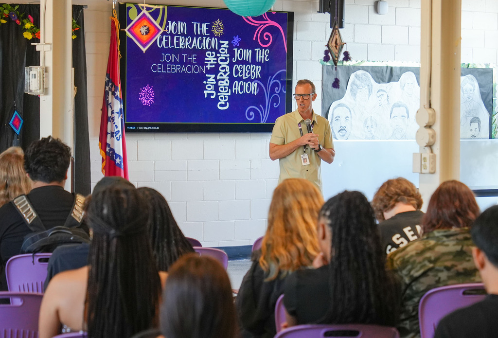 A person speaks at a microphone in front of a screen in a classroom with many students.