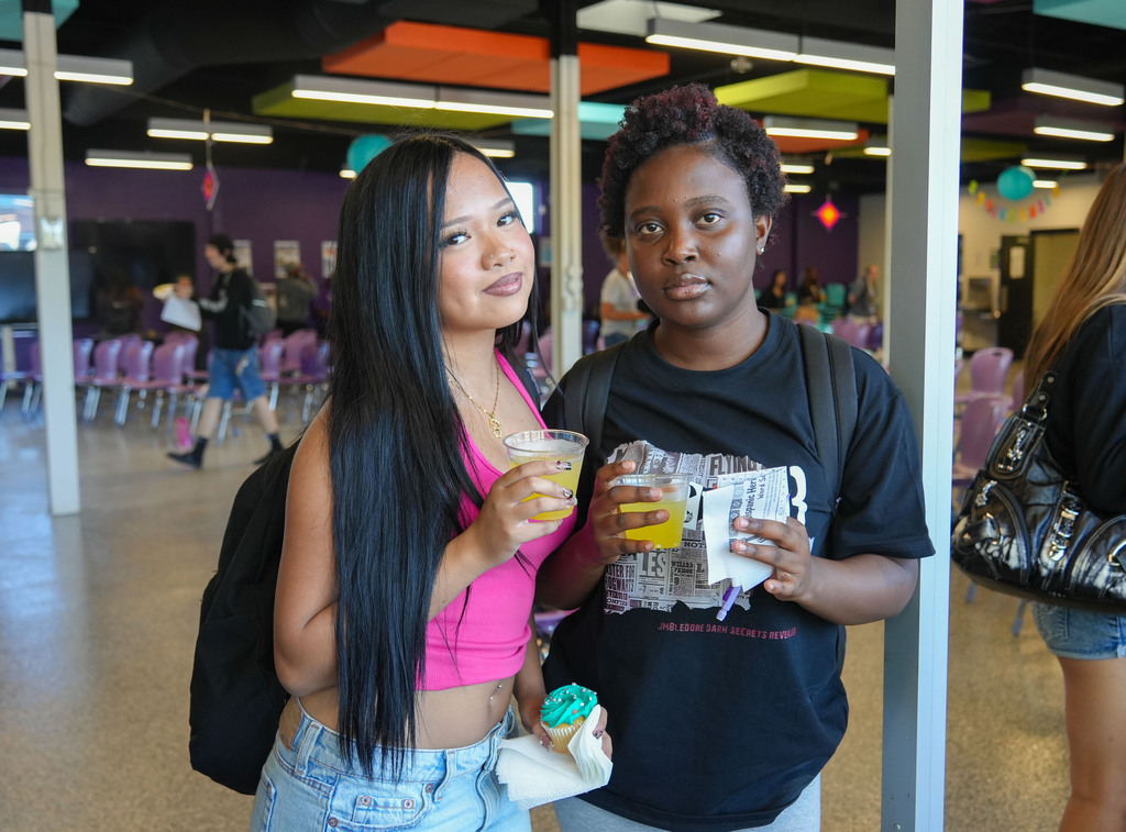 Two women in a room with chairs, a backpack, drinks, and balloons in the background.