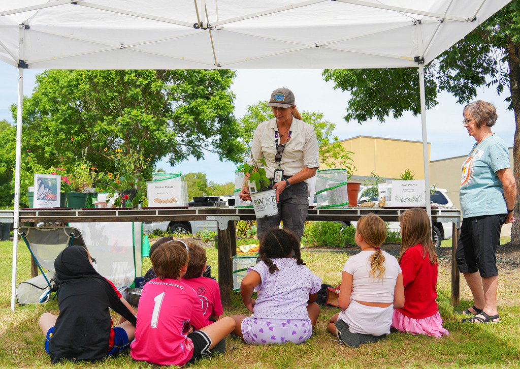 A woman stands at a table under a tent, addressing a group of children sitting on the grass.