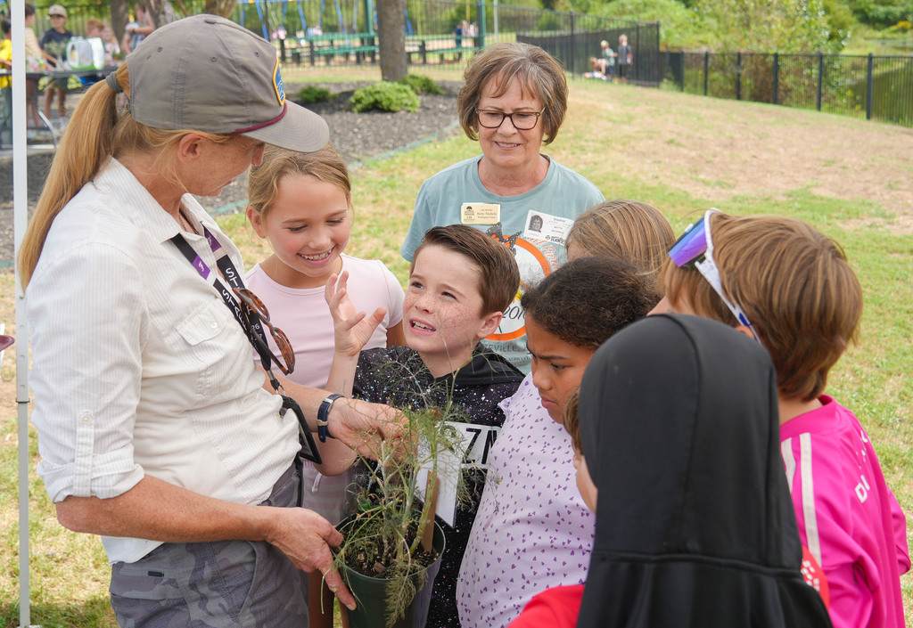 Woman with a plant speaks to a group of children, one holding a small potted plant.