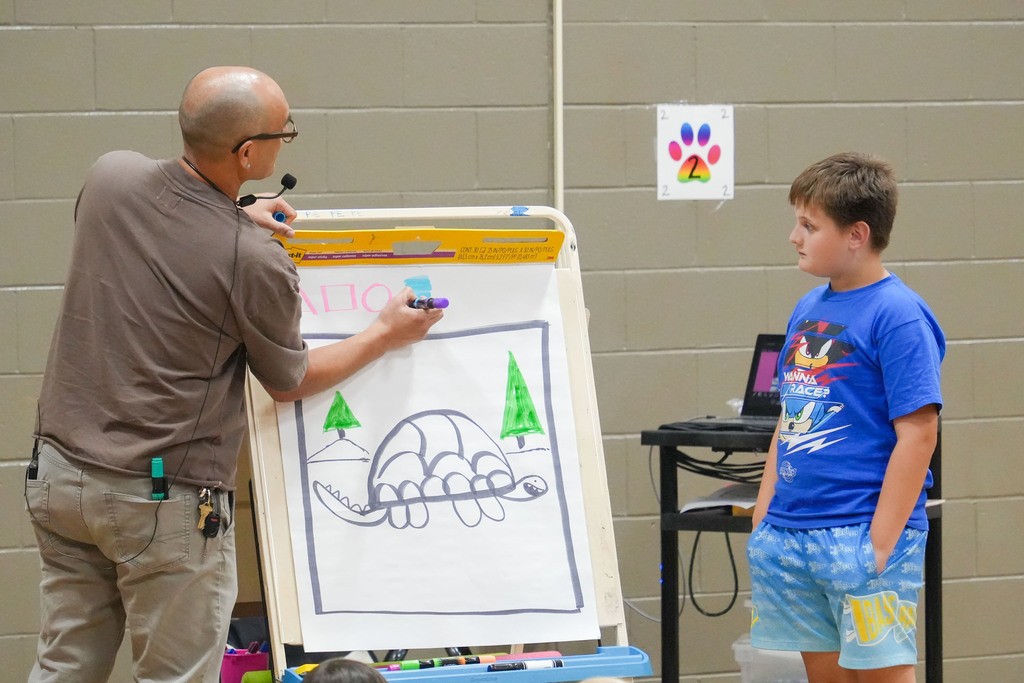 An adult draws on a whiteboard while a child watches. The drawing includes trees. A laptop is on a cart.