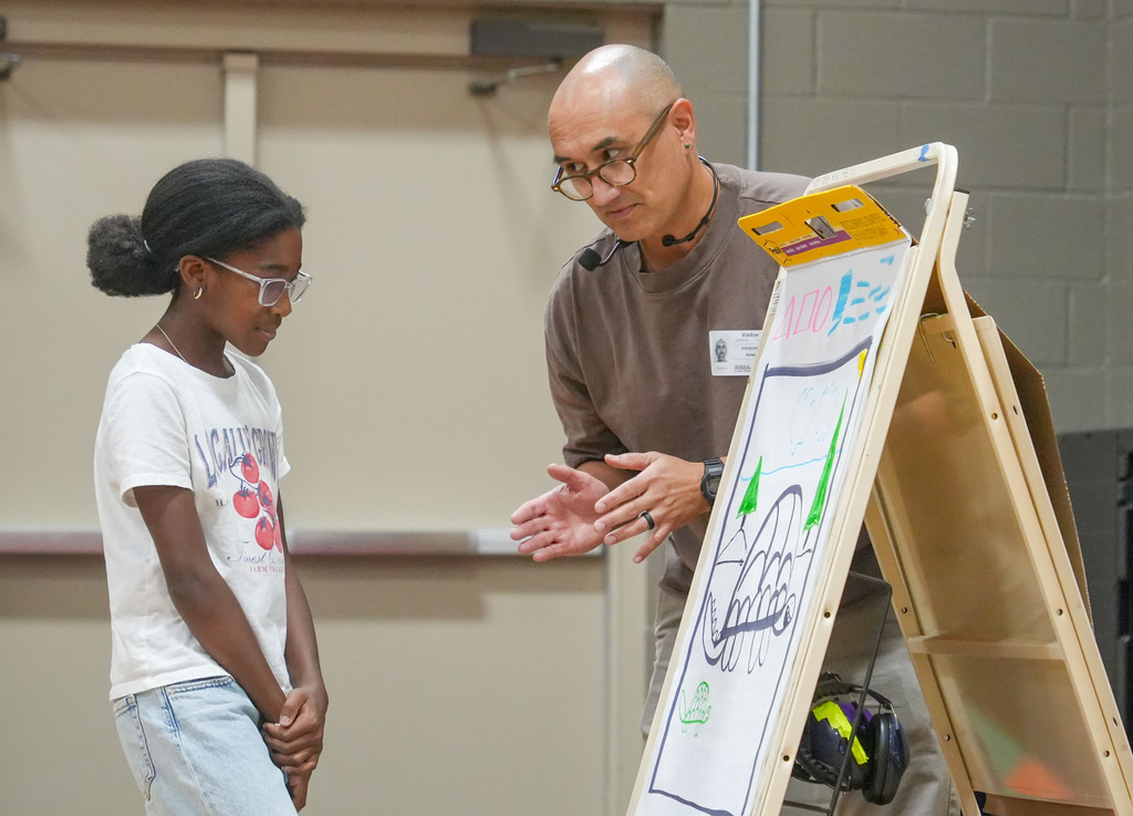 A person wearing glasses stands next to a young girl, both looking at a white drawing board.