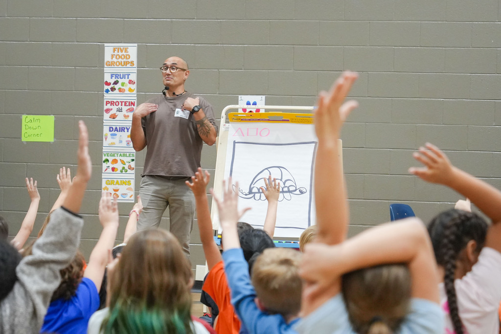 A man stands in a classroom with raised hands, addressing students. The room has a whiteboard and posters in the background.