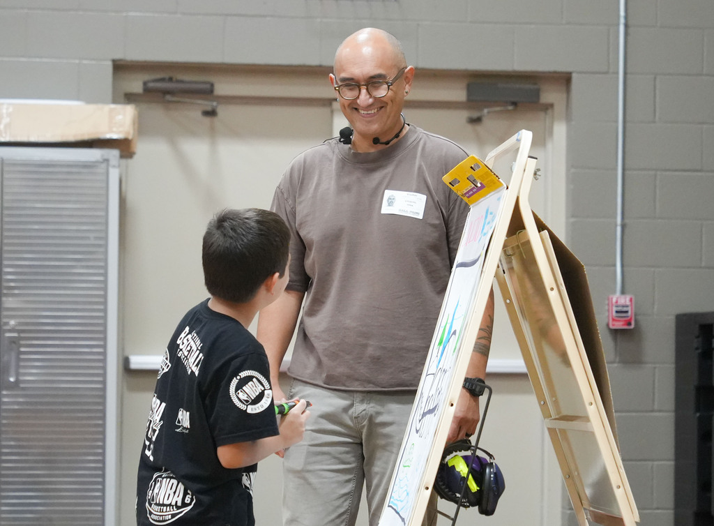 A man and a child stand before a wooden easel with a large sheet of paper. The man wears glasses and a microphone.