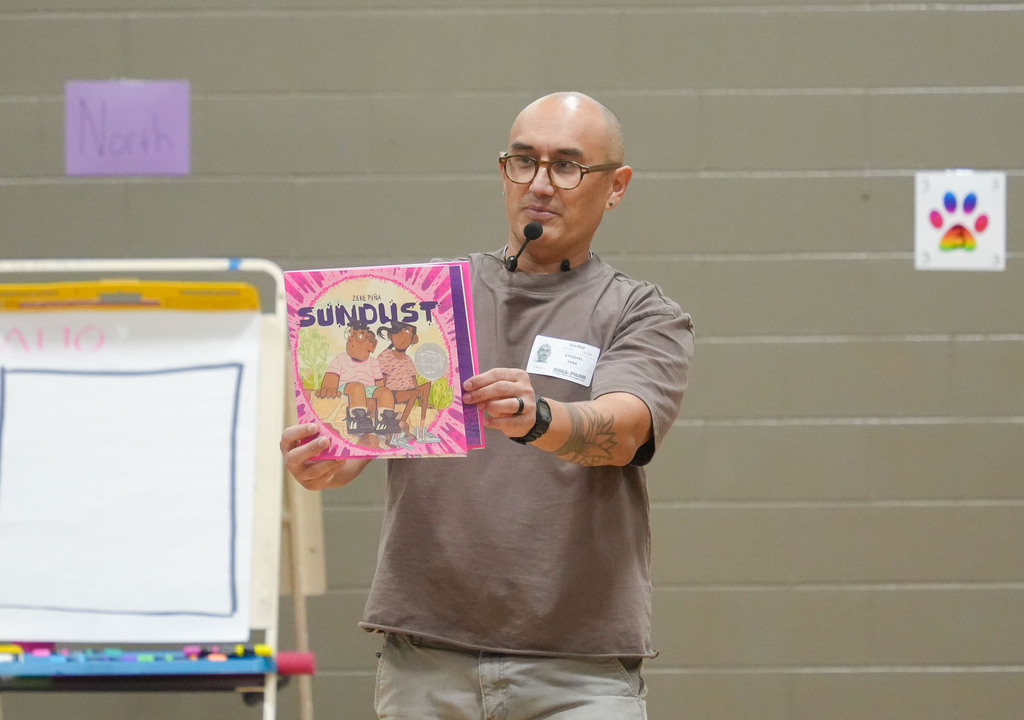 A man holding a book titled "Sunflower" speaks into a microphone. A whiteboard is to his left.