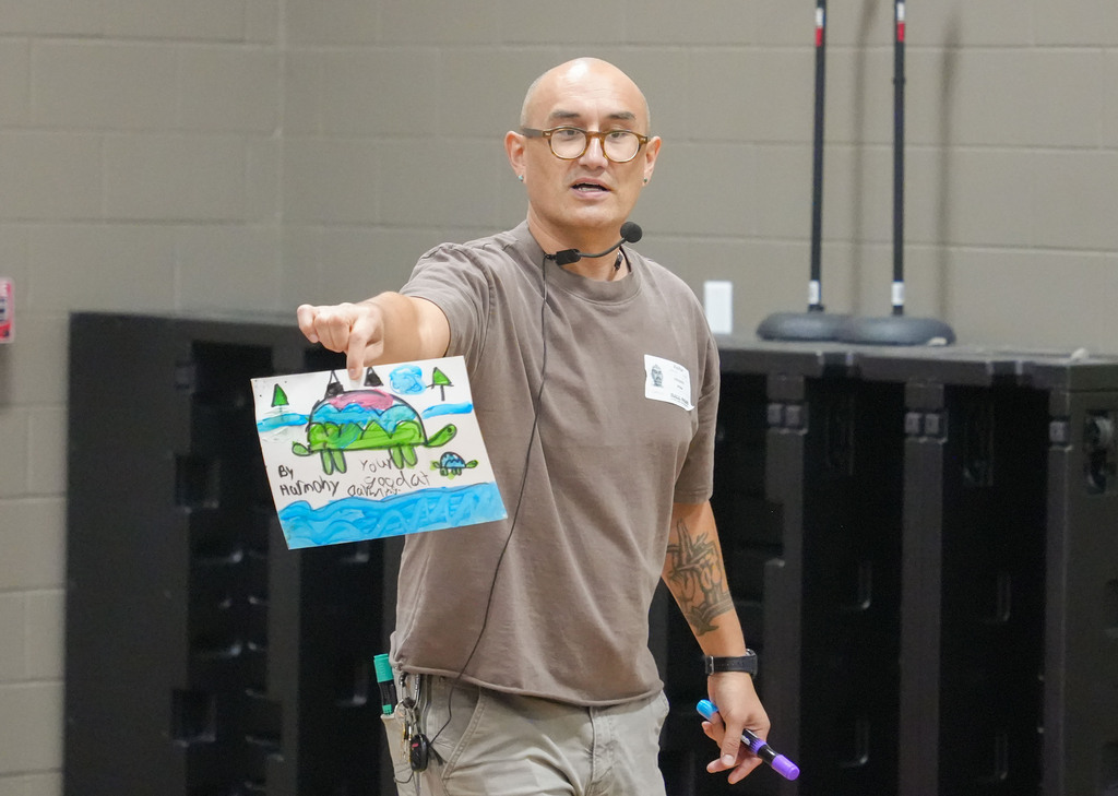 A man with glasses in a gray shirt holds a card with a drawing while gesturing.