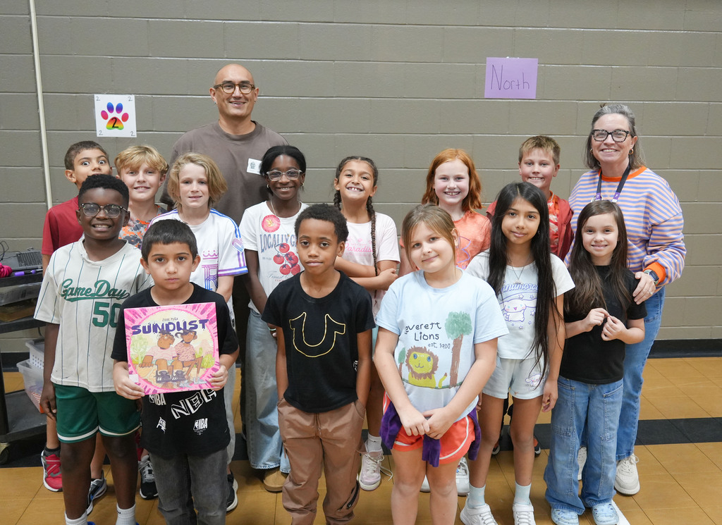 A group of children and adults stand in a room, smiling. Some hold colorful items. A white wall is in the background.