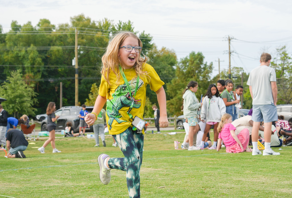A young girl wearing glasses runs on a grassy field with others in the background.
