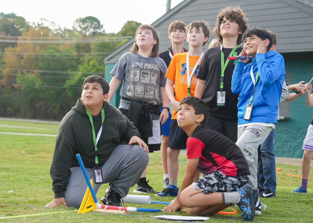 Children in a grassy area, some kneeling and others standing, wearing ID lanyards. One holds a stick.