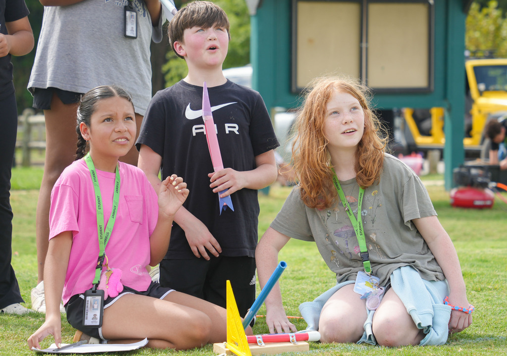 Three children on grass. One girl in pink looks sideways, another in gray holds a stick, and a boy in black looks upward.