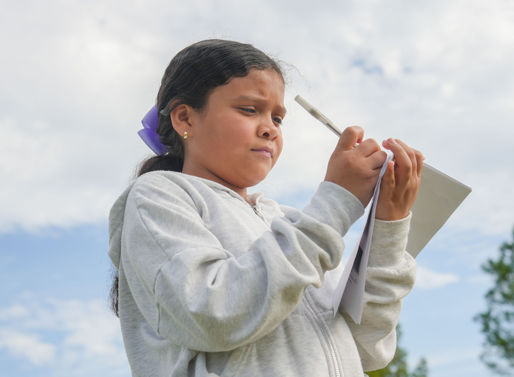 A young girl writes on a piece of paper with a pen. She wears a gray hoodie and a purple hair tie. The sky is partly cloudy.